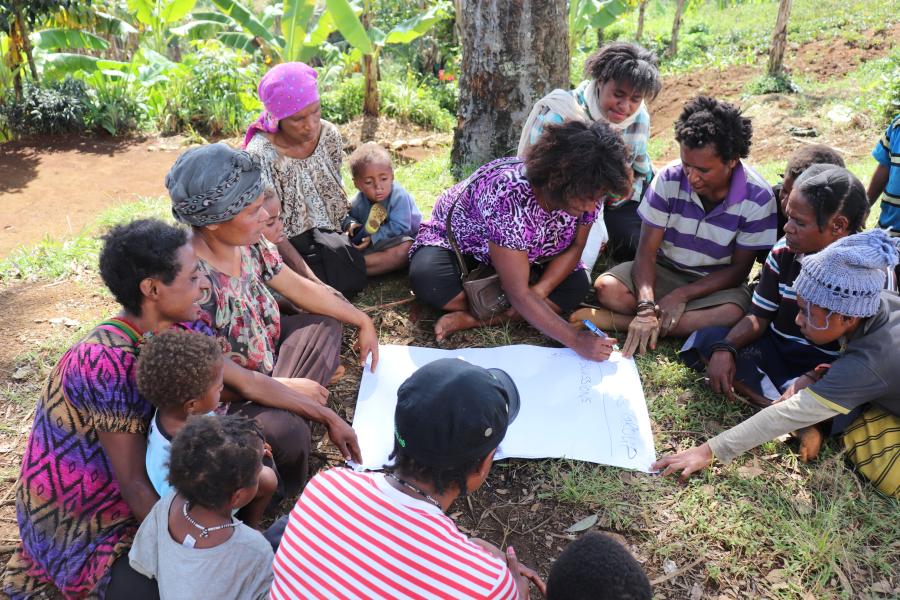 Group of people sit on the ground around a person writing on paper.